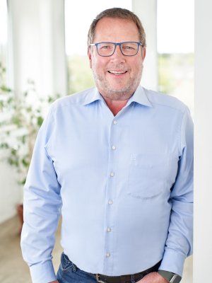 Porträt von Detlef Prien, Senior Advisor, lächelnd in einem blauen Hemd vor einem hellen, neutralen Hintergrund.