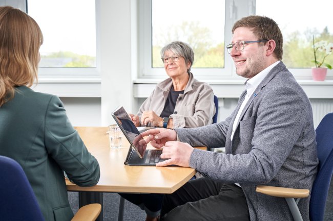 Drei Personen sitzen in einem hellen Büro an einem runden Besprechungstisch. Im Vordergrund blickt ein Mann mit Brille auf ein Tablet, während die Gruppe konzentriert und freundlich miteinander kommuniziert.