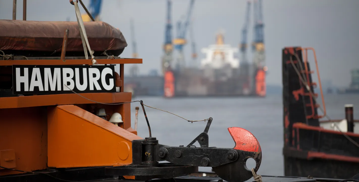 Nahaufnahme des Hecks eines orangefarbenen Schiffes mit der Aufschrift „HAMBURG“ und einem massiven schwarzen Schlepphaken im Hamburger Hafen.