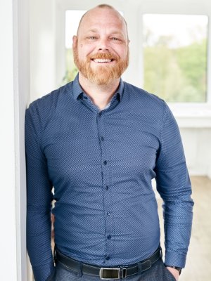 Porträt von Christian Dittmer, Head of Technical DSC, lächelnd mit Bart und blauem gemustertem Hemd vor hellem Hintergrund.