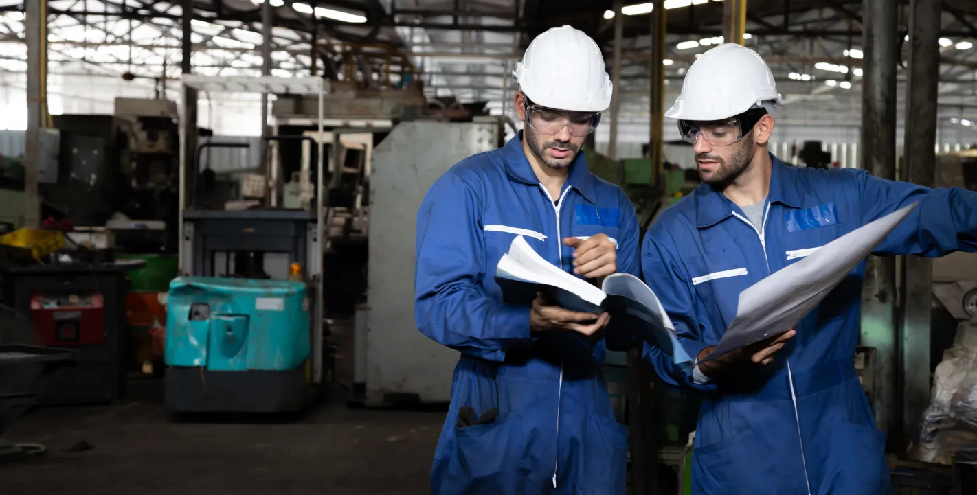 Zwei Techniker in blauen Overalls, weißen Schutzhelmen und Sicherheitsbrillen stehen in einer Industriehalle und gehen gemeinsam konzentriert technische Unterlagen durch.