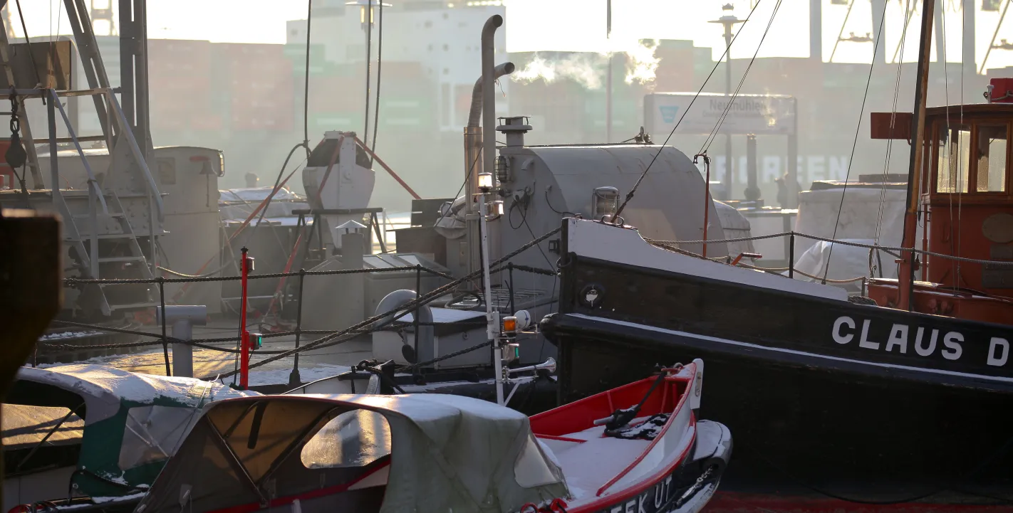 Historische Boote „Stek Ut“ und „Claus D.“ liegen im Hamburger Museumshafen Oevelgönne im winterlichen Morgenlicht vor der Kulisse von Containerbrücken.