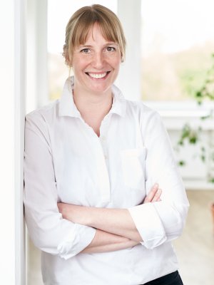 Porträtaufnahme von Annika Lohwasser - Marketing & Communications: Eine junge Frau mit blondem Haar und Pony lächelt herzlich in die Kamera. Sie trägt eine weiße Bluse, hat die Arme vor der Brust verschränkt und lehnt entspannt an einem weißen Türrahmen.