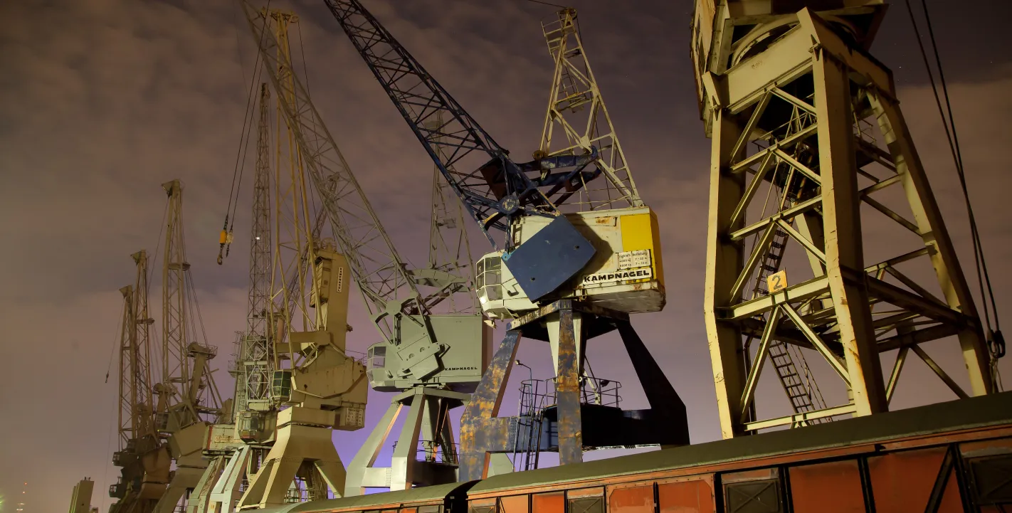 Historische Hafenkräne im Hamburger Hafen bei Nacht; im Vordergrund stehen alte braune Güterwaggons auf Schienen.