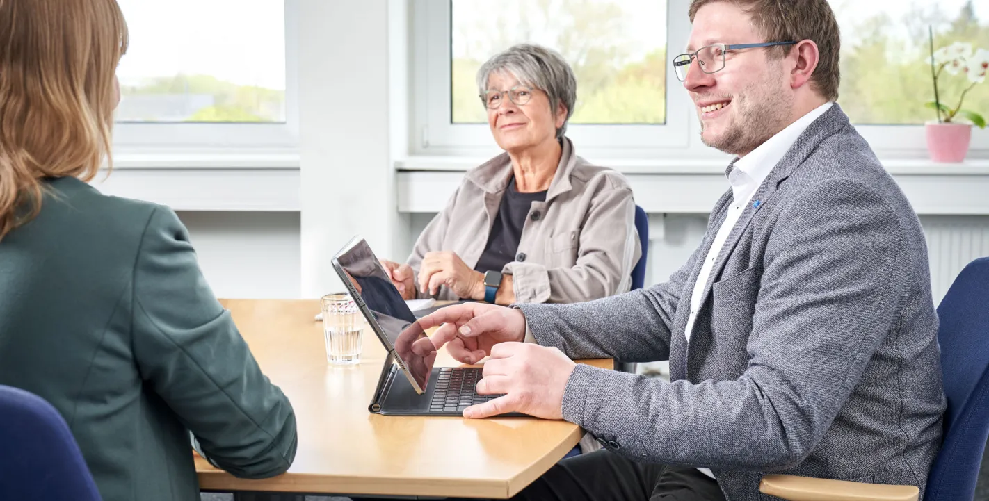 Drei Personen sitzen in einem hellen Büro an einem runden Besprechungstisch. Im Vordergrund blickt ein Mann mit Brille auf ein Tablet, während die Gruppe konzentriert und freundlich miteinander kommuniziert.