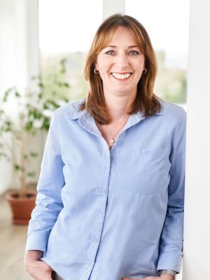 Porträtaufnahme von Melanie Urban (Finance): Eine Frau mit braunen, schulterlangen Haaren lächelt herzlich in die Kamera. Sie trägt ein hellblaues Hemd, eine dezente Halskette sowie kleine Creolen und lehnt entspannt an einer weißen Wand.