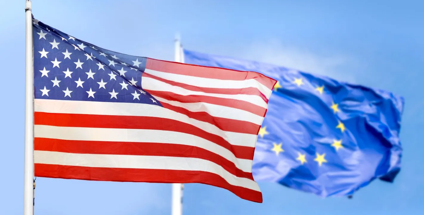 Die Flagge der USA weht im Vordergrund, dahinter ist die Flagge der Europäischen Union vor blauem Himmel zu sehen.