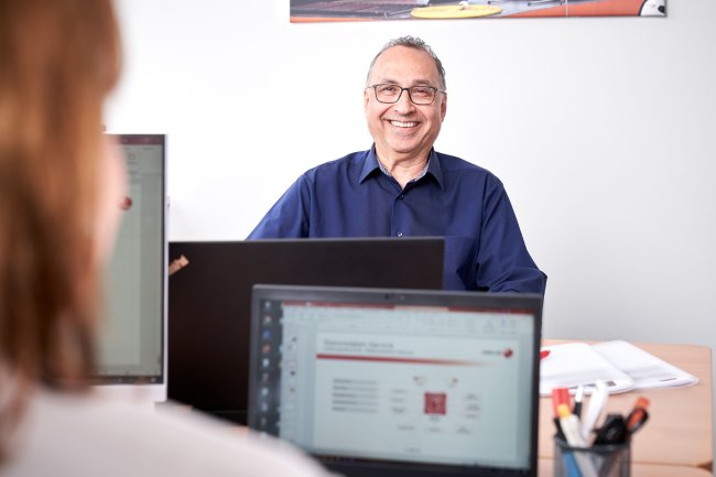 Ein lächelnder Mitarbeiter sitzt an seinem Büro-Arbeitsplatz; im Vordergrund sieht man unscharf eine Kollegin an einem Laptop.