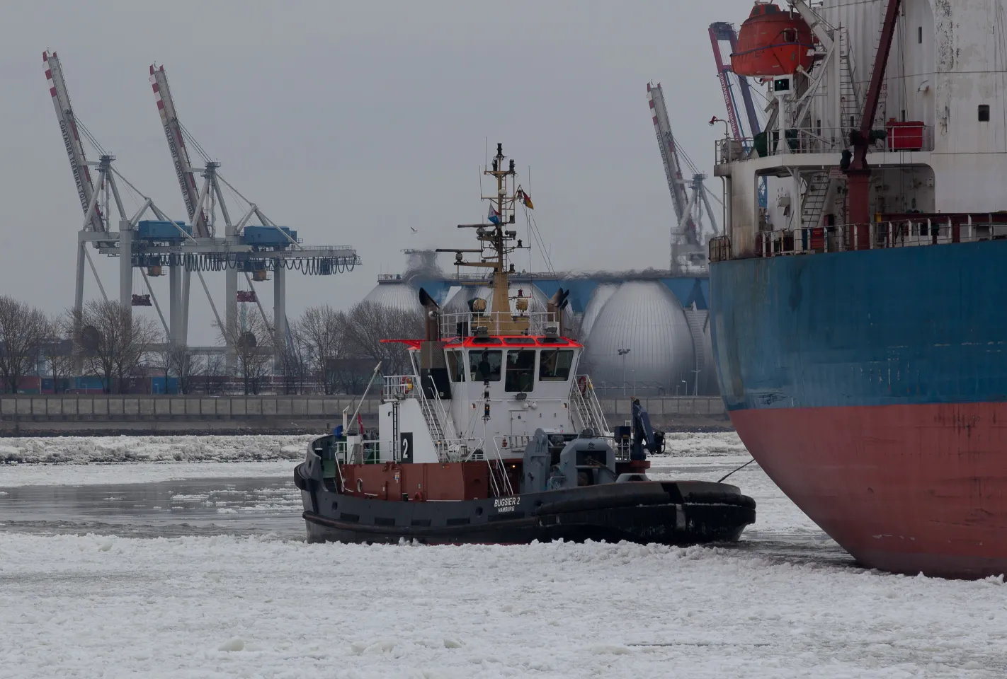Der Schlepper „Bugsier 2“ manövriert ein großes blau-rotes Schiff durch eine dichte Eisschicht im Hamburger Hafen vor der Kulisse der Köhlbrand-Gasspeicher.