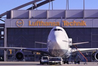 Ein großes Verkehrsflugzeug steht auf dem Rollfeld direkt vor einer Wartungshalle mit dem gelben Schriftzug „Lufthansa Technik“.
