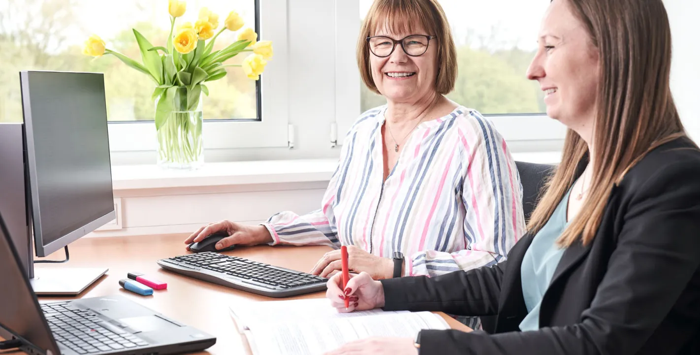 Zwei Kolleginnen arbeiten lächelnd an einem hellen Büro-Arbeitsplatz mit PC-Monitoren und einem Strauß gelber Tulpen am Fenster.