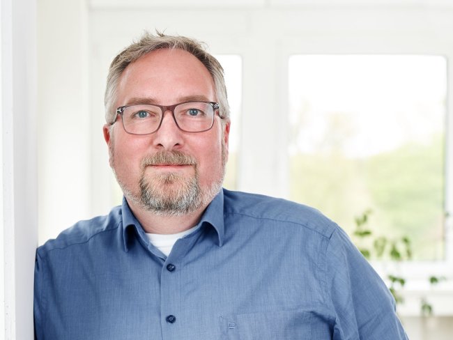 Porträt von Stephan Klein, Head of Customs & Export Service, lächelnd in einem blauen Hemd vor einem hellen Hintergrund.