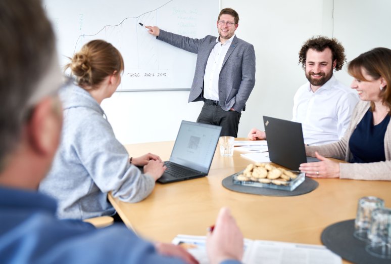 Ein Mitarbeiter hält eine Präsentation am Whiteboard vor drei Kollegen, die aufmerksam an einem Besprechungstisch sitzen.