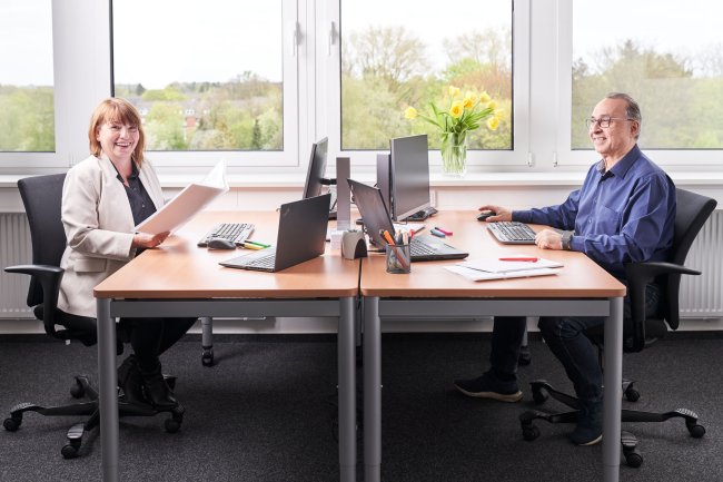 Zwei Mitarbeiter arbeiten lächelnd an einem Doppel-Schreibtisch in einem hellen Büro. Im Hintergrund ist durch ein großes Fenster grüne Vegetation und ein Strauß gelber Tulpen zu sehen.