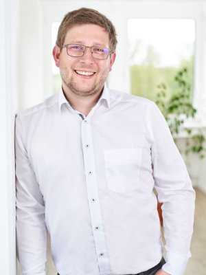 Porträt von Malte Prien (Managing Director): Ein junger Mann mit Brille und kurzem Bart lächelt herzlich. Er trägt ein weißes Hemd und lehnt entspannt an einer weißen Wand im Büro.
