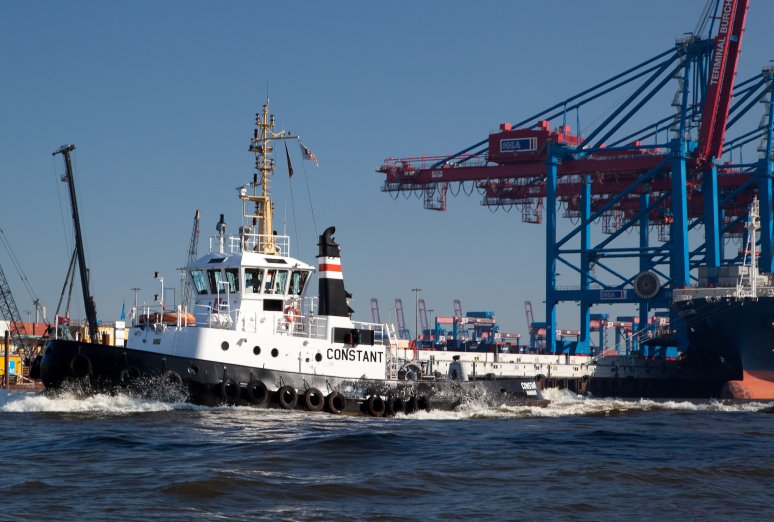 Der schwarz-weiße Schlepper „Constant“ fährt bei sonnigem Wetter im Hamburger Hafen vor den blauen Kränen des Terminals Burchardkai.