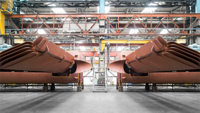 Industriehalle mit zwei riesigen, rostfarbenen Metallkonstruktionen im Vordergrund. Im Hintergrund sind Stahlträger und eine gelbe Krananlage sichtbar.
