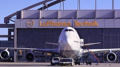 Ein großes Verkehrsflugzeug steht auf dem Rollfeld direkt vor einer Wartungshalle mit dem gelben Schriftzug „Lufthansa Technik“.